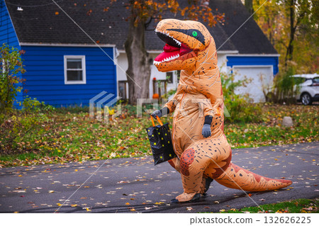 Person in inflatable dinosaur costume walking along suburban street with a Halloween candy bag. 132626225