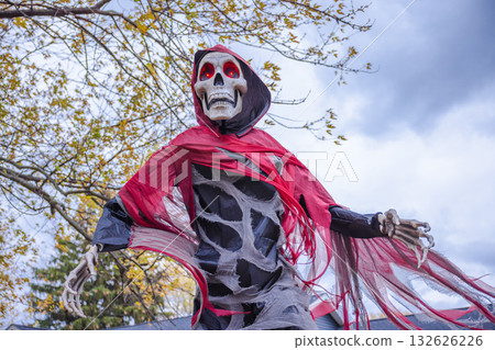 Creepy Halloween skeleton decoration with glowing red eyes and torn red cloak under cloudy autumn sky. Creepy Halloween skeleton decoration with glowing red eyes and torn red cloak under cloudy autumn sky. 132626226