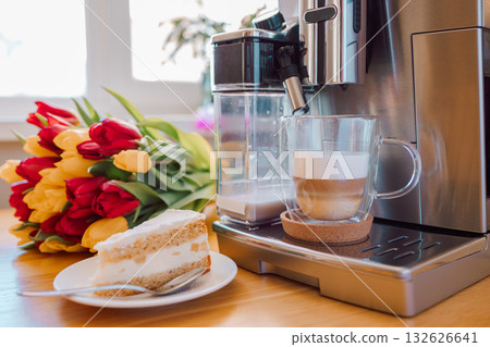 Cup of coffee with milk, piece of cake and tulips flowers on wooden kitchen table. Freshly brewed cappuccino espresso in coffee machine for breakfast. Stylish home interior decor. . High quality photo 132626641