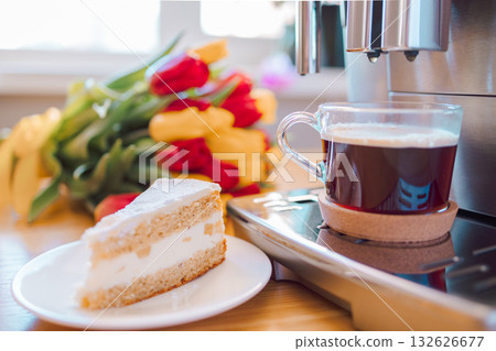 Cup of coffee with milk, piece of cake and tulips flowers on wooden kitchen table. Freshly brewed cappuccino espresso in coffee machine for breakfast. Stylish home interior decor. . High quality photo Cup of coffee with milk, piece of cake and tulips flowers on wooden kitchen table. Freshly brewed cappuccino espresso in coffee machine for breakfast. Stylish home interior decor. . High quality photo 132626677