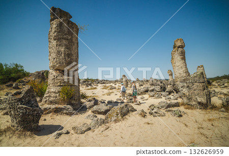 Two women walking among tall limestone columns in Pobiti Kamani, Bulgaria, a natural desert landscape with sand, rocks, and blue summer sky. 132626959