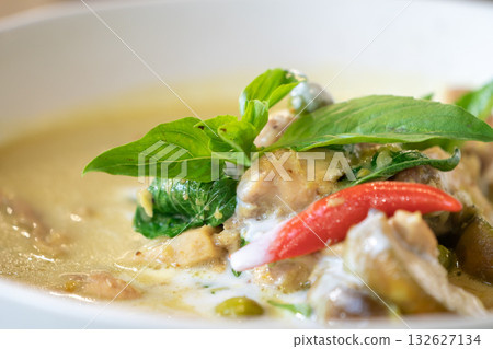 Close up to Green Curry with Chicken garnished on top with pepper and basil was served with white bowl put on wicker cream color placemat. 132627134