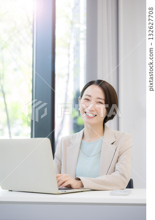 A businesswoman working on a computer by a window with greenery, looking at the camera 132627708