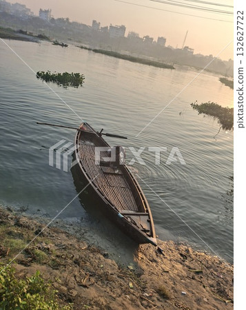 A solitary wooden boat rests on the muddy bank of a calm river at sunrise, with a distant city skyline in the background A solitary wooden boat rests on the muddy bank of a calm river at sunrise, with a distant city skyline in the background 132627722