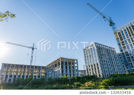 Cranes work above a busy construction site, where new residential buildings are being built alongside greenery under a clear blue sky 132633458