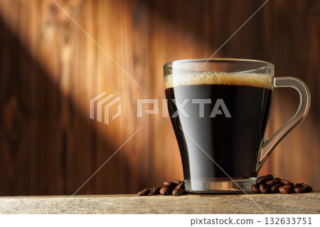 Ray of light falls on transparent cup of espresso, which sits on table with coffee beans scattered around it Ray of light falls on transparent cup of espresso, which sits on table with coffee beans scattered around it 132633751