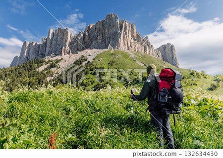 Hiker man with trekking poles, backpack and solar panel in green meadow with Bolshoy Tkhach mountain Hiker man with trekking poles, backpack and solar panel in green meadow with Bolshoy Tkhach mountain 132634430