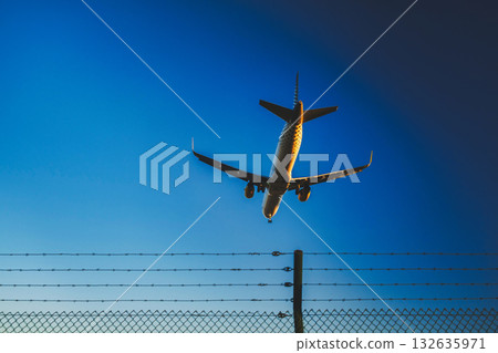 This image shows an airplane in the middle of landing or taking off, seen from a perspective where it is flying low over a perimeter fence. The fence is likely part of the security boundary for an This image shows an airplane in the middle of landing or taking off, seen from a perspective where it is flying low over a perimeter fence. The fence is likely part of the security boundary for an 132635971