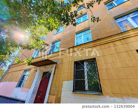 Old residential building with a yellow facade, red entrance door, and barred windows under bright sunlight filtering through tree leaves. The image combines a sense of safety and aging infrastructure 132639004