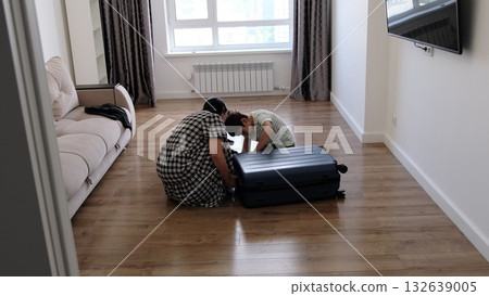 Mother and child prepare a blue suitcase on the floor of a tidy apartment, captured in soft daylight. The image symbolizes readiness, family support, and the excitement of exploring new places. Mother and child prepare a blue suitcase on the floor of a tidy apartment, captured in soft daylight. The image symbolizes readiness, family support, and the excitement of exploring new places. 132639005