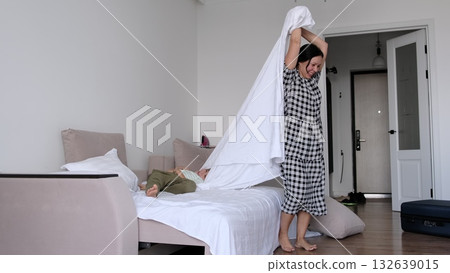 The sheet billows between mother and child as they make the bed together, bright and airy with uncluttered lines. Shared housekeeping connects generations and keeps homes healthy and welcoming. The sheet billows between mother and child as they make the bed together, bright and airy with uncluttered lines. Shared housekeeping connects generations and keeps homes healthy and welcoming. 132639015