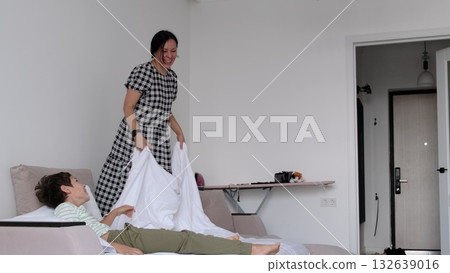 A mother laughs as she lifts a white sheet over her son on a wide bed, captured candidly with soft daylight. The moment celebrates routines that build comfort, bonding, and resilience in a real home. A mother laughs as she lifts a white sheet over her son on a wide bed, captured candidly with soft daylight. The moment celebrates routines that build comfort, bonding, and resilience in a real home. 132639016