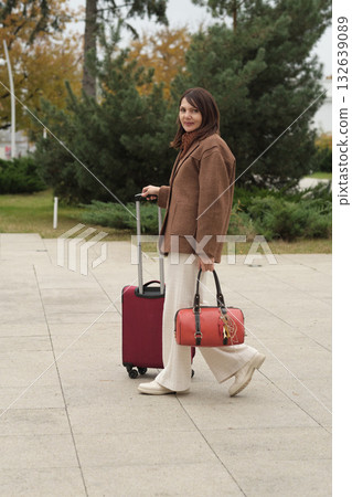 A traveler with a red suitcase and a matching handbag outdoors on a calm autumn day. This symbolizes solo journeys and the freedom to explore new places. 132639089