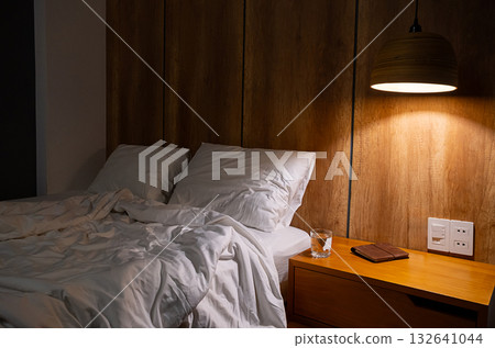 Unmade bed with white linen and pillows in hotel room with glass of water, book on nightstand at night. 132641044