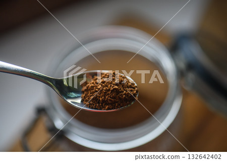 A spoon dipped into a glass jar filled with instant coffee granules on a wooden table, captured in warm natural light. A spoon dipped into a glass jar filled with instant coffee granules on a wooden table, captured in warm natural light. 132642402