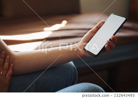Young woman hold smartphone with white screen while sitting on a couch Young woman hold smartphone with white screen while sitting on a couch 132643493