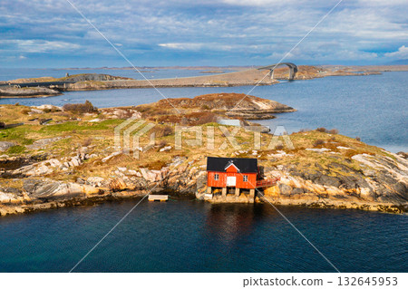 Small house nestled on rocky brown islet among expansive fjord waters of Norwegian coast. Mountains highlight dramatic natural beauty of Norway Small house nestled on rocky brown islet among expansive fjord waters of Norwegian coast. Mountains highlight dramatic natural beauty of Norway 132645953