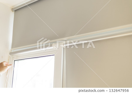 Process of installing light gray roller blinds on window indoors. 132647956