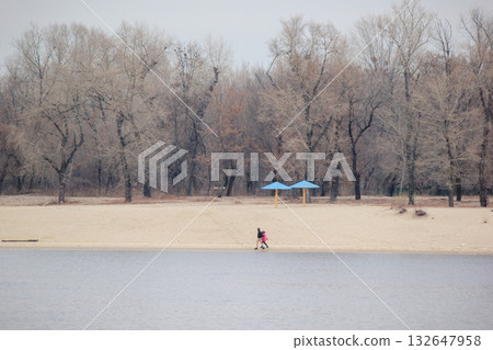 A lone figure walks on a deserted river beach with bare trees and blue umbrellas on a cold autumn day 132647958