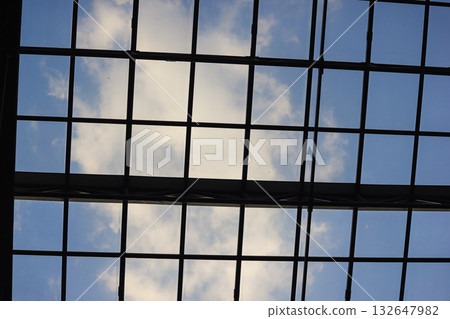 Looking up at a glass roof grid against a bright blue sky with white clouds. Abstract modern architecture 132647982