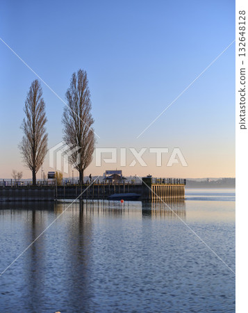 Serene morning at the lakeside dock with reflections and tall trees during sunrise Serene morning at the lakeside dock with reflections and tall trees during sunrise 132648128