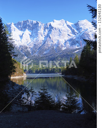 Majestic snow-capped mountains reflect in serene lake surrounded by evergreen trees at midday Majestic snow-capped mountains reflect in serene lake surrounded by evergreen trees at midday 132648180