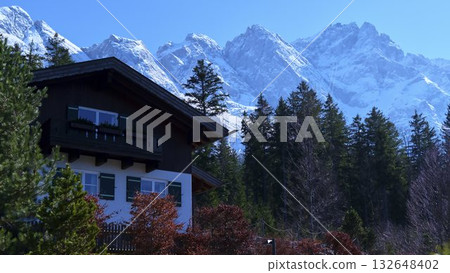 Scenic mountain view with a cozy house surrounded by trees and snow-capped peaks in a clear blue sky Scenic mountain view with a cozy house surrounded by trees and snow-capped peaks in a clear blue sky 132648402