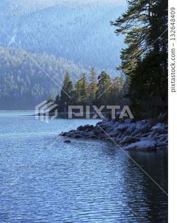 Tranquil lake view with rocky shoreline and forested mountains during early morning light Tranquil lake view with rocky shoreline and forested mountains during early morning light 132648409