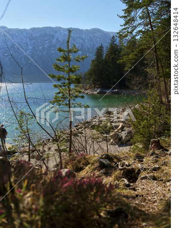 Scenic lakeside view with a hiker exploring rocky shoreline on a clear day surrounded by mountains Scenic lakeside view with a hiker exploring rocky shoreline on a clear day surrounded by mountains 132648414