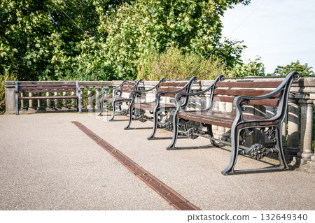 A row of classic wooden benches with ornate black iron frames stands neatly aligned beside a stone A row of classic wooden benches with ornate black iron frames stands neatly aligned beside a stone 132649340