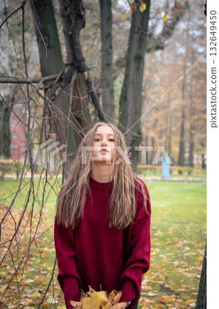 Teenage girl in a red sweater stands in a park holding autumn leaves, surrounded by trees and soft natural light 132649450