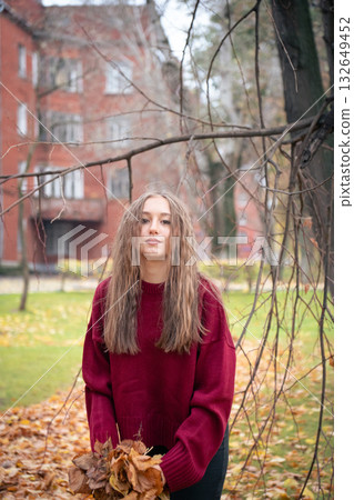 Teenage girl in a red sweater stands in a park holding autumn leaves, with soft light and warm tones creating a serene mood Teenage girl in a red sweater stands in a park holding autumn leaves, with soft light and warm tones creating a serene mood 132649452
