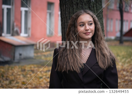 Young woman in a dark blazer and mesh top smiles softly while standing by a tree in an autumn park Young woman in a dark blazer and mesh top smiles softly while standing by a tree in an autumn park 132649455