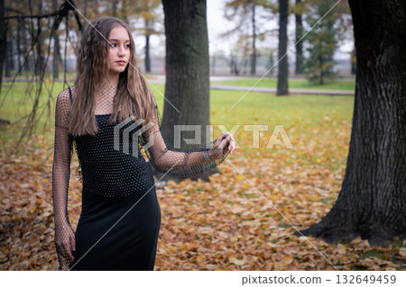 Young woman in a black mesh top stands among autumn trees and fallen leaves, looking away with a calm and thoughtful expression 132649459