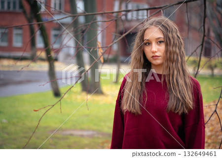 Teenage girl in a red sweater stands outdoors in soft autumn light, with blurred brick buildings and green grass in the background Teenage girl in a red sweater stands outdoors in soft autumn light, with blurred brick buildings and green grass in the background 132649461