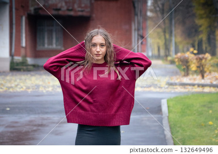 Teenage girl in a red oversized sweater stands on a wet autumn street, holding her hair with both hands and looking confidently at the camera 132649496