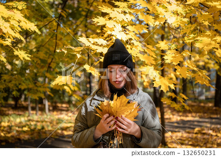 Young woman in a dark blazer and mesh top smiles softly while standing by a tree in an autumn park 132650231