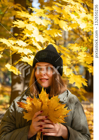Woman in a green jacket and black hat holds yellow maple leaves while looking up among bright autumn foliage Woman in a green jacket and black hat holds yellow maple leaves while looking up among bright autumn foliage 132650236