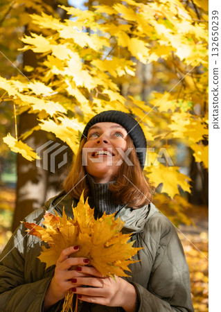 Woman in a green jacket and black hat holds yellow maple leaves while looking up among bright autumn foliage Woman in a green jacket and black hat holds yellow maple leaves while looking up among bright autumn foliage 132650239