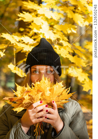 Woman in a black hat and green jacket holds bright yellow maple leaves in front of her face, surrounded by golden autumn foliage Woman in a black hat and green jacket holds bright yellow maple leaves in front of her face, surrounded by golden autumn foliage 132650248