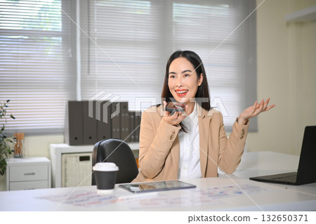 Smiling businesswoman talking on smartphone using speakerphone in office, expressing positivity Smiling businesswoman talking on smartphone using speakerphone in office, expressing positivity 132650371