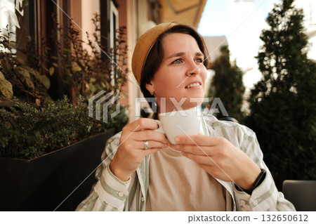 Young woman in yellow cap holding a cup of coffee and looking away while sitting at outdoor cafe. Good morning in city concept 132650612