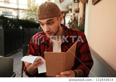 Shocked man holding a restaurant bill and menu while sitting at outdoor cafe 132650614
