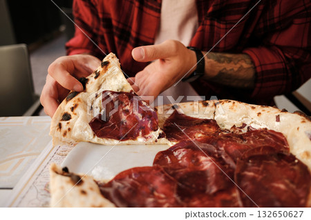 Close up of a man in a red checkered shirt enjoying pizza with cured meat at an outdoor cafe Close up of a man in a red checkered shirt enjoying pizza with cured meat at an outdoor cafe 132650627
