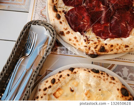 Freshly baked two pizza with golden cheese next to basket of cutlery, ready for perfect dining experience. Italian outdoor cafe, close view 132650628