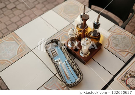 Neatly arranged dining table with olive oil bottles, condiments, and metal cutlery in wicker basket Neatly arranged dining table with olive oil bottles, condiments, and metal cutlery in wicker basket 132650637