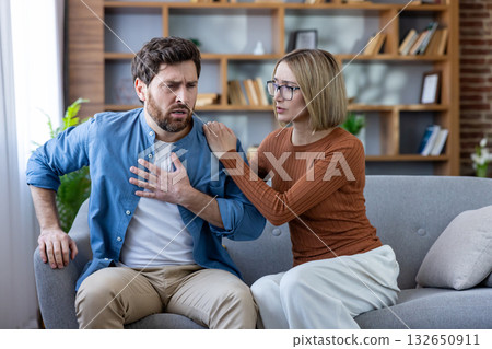 Man experiencing acute chest pain and shortness of breath while sitting on a couch at home, receiving concerned support and comfort from a woman during a health emergency or panic attack 132650911
