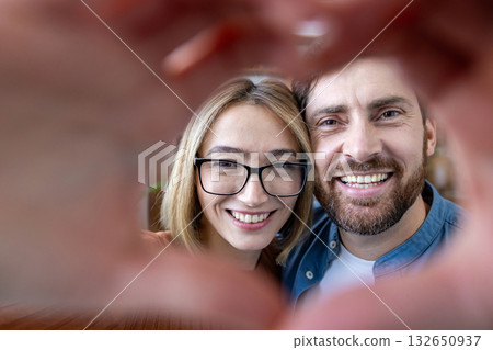 Happy diverse couple making a heart with their hands around their faces, smiling at the camera in a warm, candid indoor portrait that radiates love and togetherness 132650937