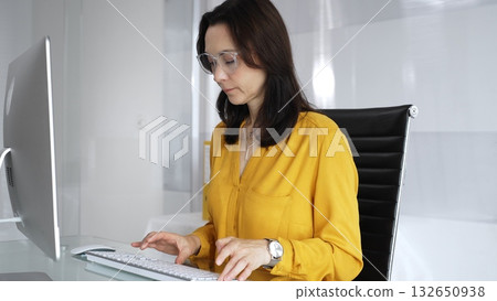 Professional woman wearing yellow casual blouse and glasses focused on laptop screen at office desk, typing and analyzing data for business tasks. Audit in business 132650938