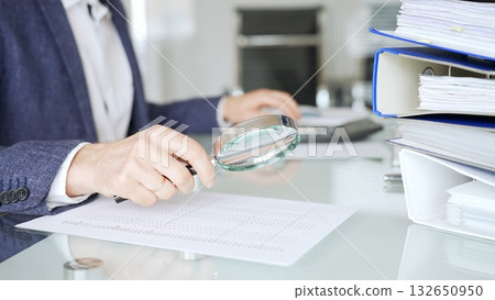Accountant hand holding magnifying glass checking financial document numbers on office desk, analyzing taxes and data. Audit and taxes in business 132650950
